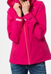 Persona che indossa una giacca a cappuccio fucsia brillante con zip frontale e cinturini in Velcro sui polsi, abbinata a jeans blu, mani vicino al colletto.