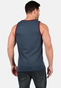 Camiseta sin mangas azul hecha de una mezcla de algodón, con un ribete en color marrón granate en las sisas y el escote. La tela tiene una textura suave y un corte casual.