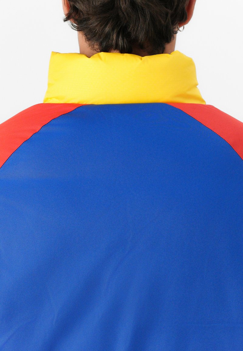 Osoba widziana z tyłu, ubrana w kurtkę w kolorze bloków z żółtym kołnierzem, niebieską plecą i czerwonymi panelami na ramionach na białym tle.