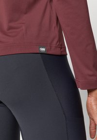 Bordo sportlik pikkade varrukatega topp logotagiga, koos sobivate mustade retuusidega. Pehme, niiskust eemal hoidav kangas, sile tekstuur.