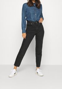 Mörk tvättade högmidjade jeans med rak bendesign, parat med en ljusblå denimskjorta med knappar. Vita sneakers fulländar looken.