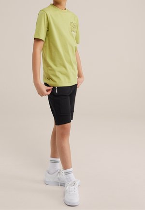 Młoda osoba ubrana w jasnozieloną koszulkę z krótkim rękawem, czarne szorty z boczną kieszenią, białe skarpetki za kostkę z szarymi paskami oraz białe sneakersy stoi.