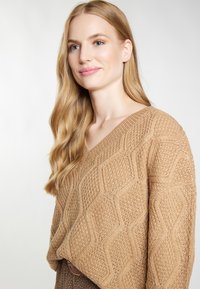 Sweter w splot wzoru plastra miodu z dekoltem w serek w jasnobrązowym kolorze, o teksturowym wykończeniu i luźnym kroju, noszony na wzorzystym elemencie odzieży.