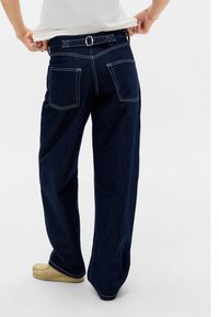 Tamno plave širom krojene traperice s pojasom, koje imaju dva stražnja džepa i kontrastne bijele šavove. Glatka denim tekstura, ležeran dizajn.