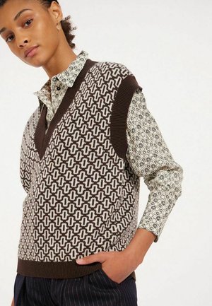 Gilet en tricot marron avec un motif géométrique blanc et un décolleté en V, superposé sur une chemise à motifs clairs à manches longues.