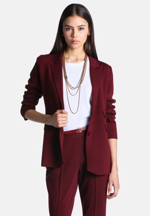 Femme aux longs cheveux foncés portant un costume bordeaux, un haut blanc, un collier doré superposé et des boucles d'oreilles dorées, debout avec une main sur la veste.