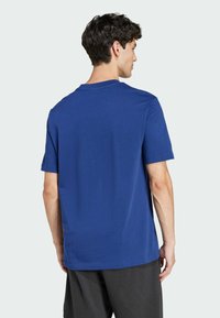 Modra bombažna majica s kratkimi rokavi, okroglo izrezana in gladke teksture, prikazana s hrbtne strani, v kombinaciji s črnimi kratkimi hlačami.