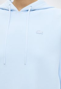 Sweat à capuche bleu clair avec cordons de serrage et petit logo crocodile texturé sur la poitrine gauche.