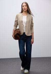 Blazer texturé beige, haut blanc, jeans larges bleu foncé, portant un sac en suède marron, avec des baskets blanches, sur un fond neutre.