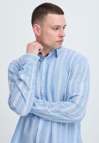 Chemise boutonnée bleu clair à rayures avec une coupe décontractée. Tissu doux, manches longues et petits boutons, avec des rayures blanches verticales.