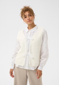 Biały dziergany bezrękawnik z dekoltem w serek, o miękkiej fakturze, z przezroczystymi wiązaniami z wstążki na środku, noszony na białej wzorzystej koszuli.