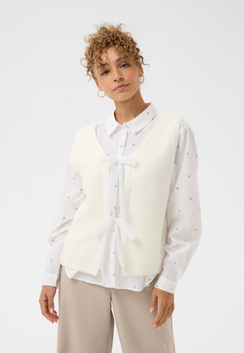 Biały dziergany bezrękawnik z dekoltem w serek, o miękkiej fakturze, z przezroczystymi wiązaniami z wstążki na środku, noszony na białej wzorzystej koszuli.