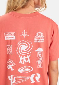 Koraalkleurig katoenen t-shirt met verschillende witte grafische prints, waaronder logo's en abstracte ontwerpen, op de achterkant. Korte mouwen met een ronde hals.