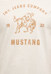 Graphique d'un cavalier sur un cheval cabré avec "INT. JEANS COMPANY 1932" au-dessus et "MUSTANG" en gras en dessous sur un fond de tissu blanc.