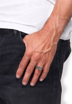 Silver metal ring with engraved Roman numerals, worn on the index finger. Hand rests on dark jeans, showcasing skin texture and details.