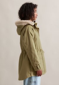 Vrouw met krullend haar die een olijfgroen parka met een beige fleece-gevoerde capuchon draagt, staande opzij tegen een effen achtergrond.