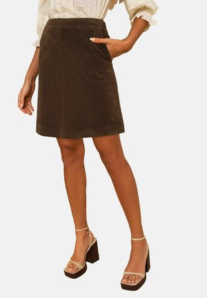 Femeie purtând o fustă maro închis până la genunchi cu buzunare, sandale bej cu toc înalt și o bluză cu mâneci lungi de culoare deschisă.