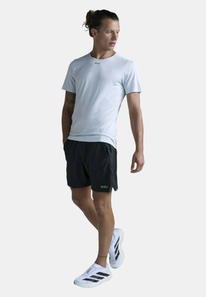 Mand iført lys grå t-shirt, sorte shorts og hvide sneakers, stående med hænderne i lommen og kigger til siden.