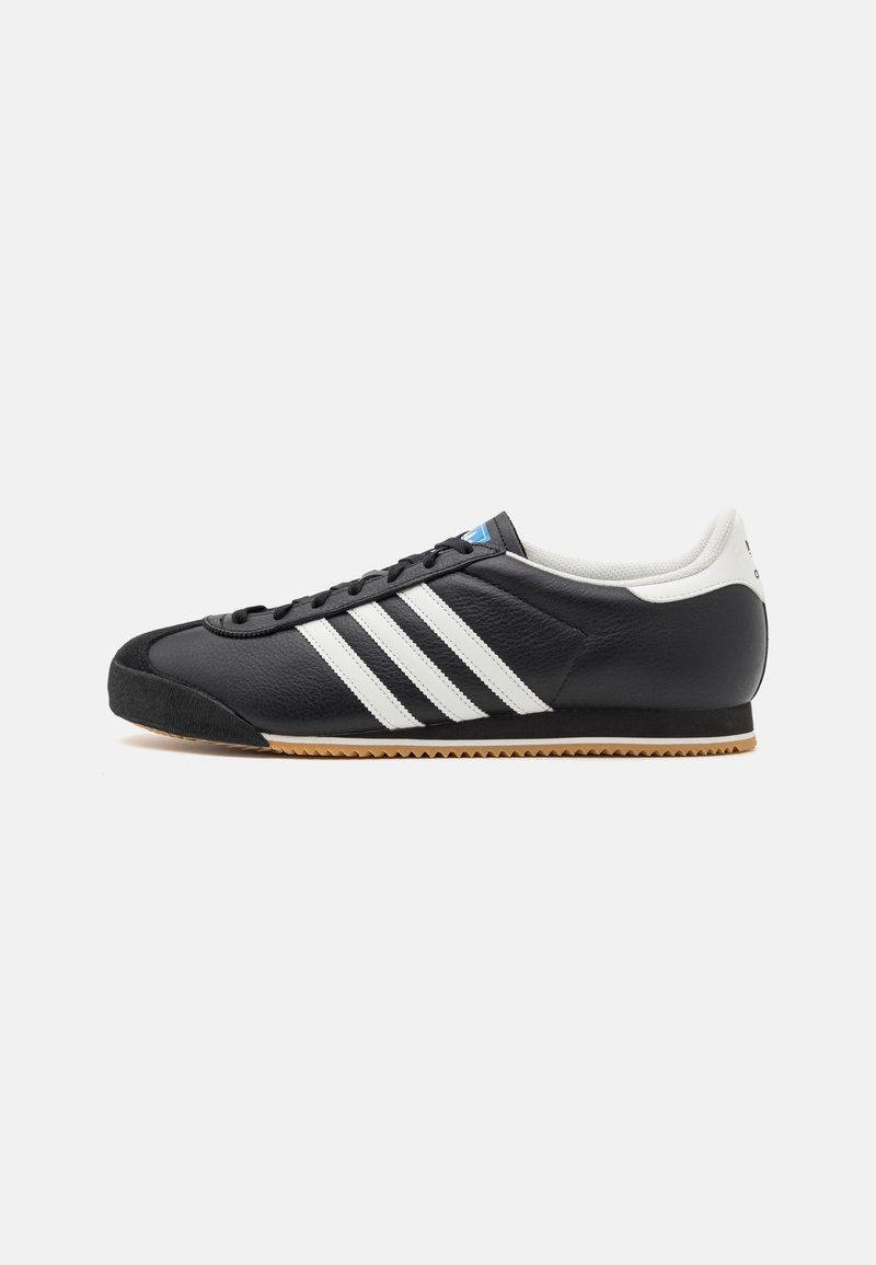 Schwarze Sportschuhe mit drei weißen Streifen, Obermaterial aus Leder, Gummisohle, geschwungene Form und strukturierte Oberfläche. Blauer Akzent auf der Zunge.