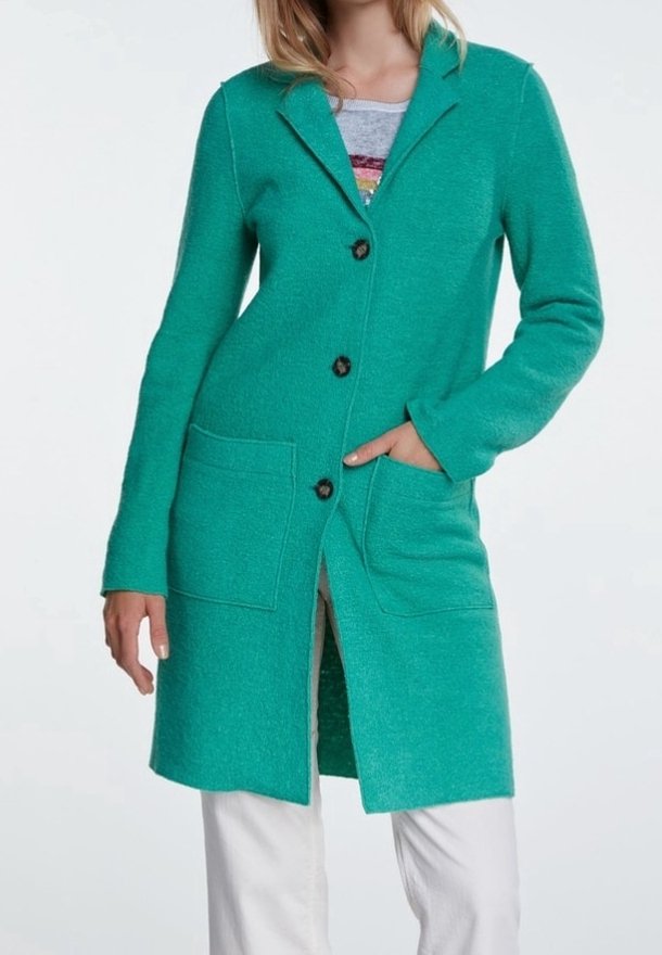 Femme portant un long cardigan bleu-vert avec de grandes poches avant, sur un haut gris et un pantalon blanc, main dans la poche.