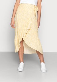 Gonna avvolgente gialla a fiori con orlo arricciato, realizzata in tessuto leggero. Caratterizzata da un fiocco in vita e abbinata a sneakers bianche.