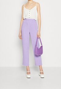 Haut blanc sans manches avec boutons, pantalons larges violets clairs, et sac à main violet ; le mannequin porte des chaussures blanches à plateforme.