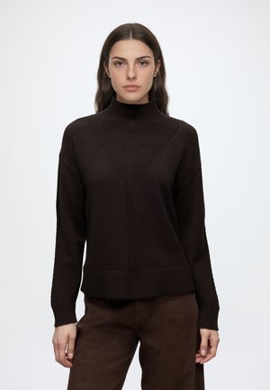 Ung kvinde med langt brunt hår iført mørkebrunt ribbet sweatertøj og matchende bukser, vendt front mod en ensartet lys baggrund.