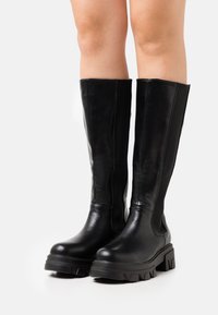 Botas negras de cuero hasta la rodilla con suela de goma gruesa, paneles elásticos laterales y detalles de costura en contraste. Textura suave con un diseño elegante.