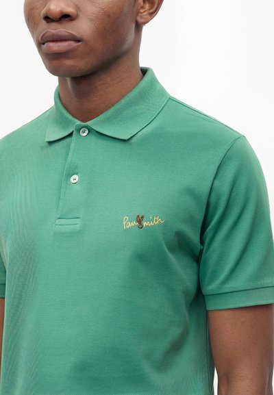 Homme portant un polo vert à manches courtes avec un petit logo brodé sur la poitrine, debout devant un fond blanc.