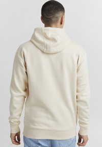 Felpa color crema con cappuccio regolabile, polsini e orlo a costine. Tessuto morbido, senza motivi, finitura liscia, abbinata a jeans.