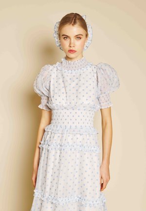 Tânără purtând o rochie albă cu gulere înalte, mâneci bufante și buline albastre, cu o bentită asortată cu volane, stând în fața unui fundal bej.