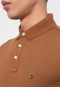 Tuvumā redzams cilvēks, kurš valkā brūnu polo kreklu ar mazu sarkanu, baltu un zilu taisnstūrveida logotipu uz krūts un trim baltām pogām uz apģērba priekšpuses.