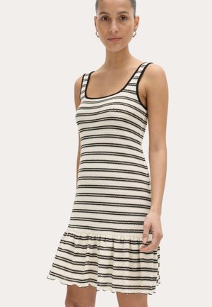 Mujer con vestido sin mangas de rayas horizontales en blanco y negro con bajo con volantes, de pie contra un fondo claro y liso.