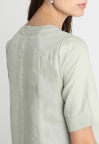 Blouse en lin vert clair à manches courtes, avec un col rond et une fermeture éclair à l'arrière. Texture lisse et coupe décontractée.