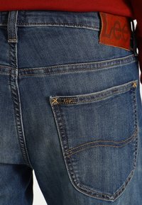 Jeans de denim azul con una etiqueta de cuero que presenta el logo "Lee". Costuras distintivas en los bolsillos traseros, ajuste regular, tela ligeramente desgastada.
