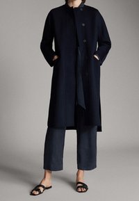 Cappotto di lana navy con colletto alto, bottoni frontali e tasche laterali, abbinato a pantaloni scuri e sandali neri. Texture liscia, design minimal.