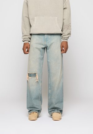 Person iført beige hættetrøje, lyseblå jeans med vide ben og et revet venstre knæ samt beige sko, stående mod en ensfarvet baggrund.