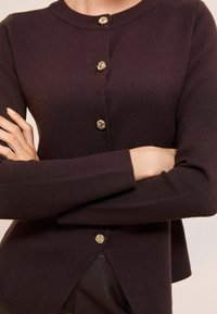 Tumši purpursarkans kardigans ar apaļu apkakli, kuram ir četri dekoratīvi zelta pogas un izsmalcināta tekstūra. Ar nedaudz izliekta apakšmalu.