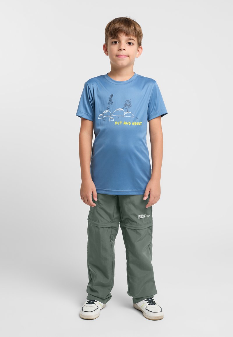 Blaues T-Shirt mit kurzen Ärmeln und graphic Print, kombiniert mit grünen Cargo-Hosen mit Taschen und einem Logo. Schlichte weiße Schuhe runden den Look ab.