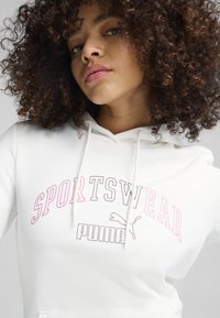 Sudadera blanca hecha de material suave con un bolsillo tipo canguro en el frente. Presenta la palabra "SPORTSWEAR" en letras en negrita rosa y gris, y un logo de Puma.