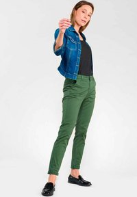 Veste en jean bleu foncé, haut noir et pantalon vert. Les pantalons présentent une coupe ajustée, une texture lisse et des ourlets retroussés ; les chaussures sont des mocassins noirs et brillants.