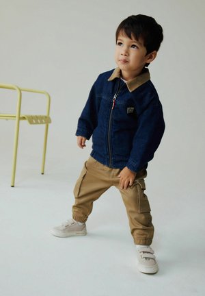 Giacca di jeans con colletto color cuoio, pantaloni cargo beige e scarpe chiare. La giacca presenta una zip frontale e una toppa con logo.