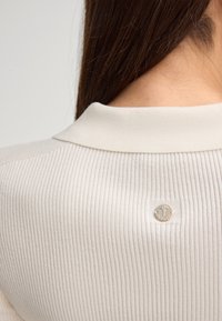 Primo piano del collo sul retro di un maglione a coste crema con un piccolo tag in metallo rotondo attaccato sotto il colletto.