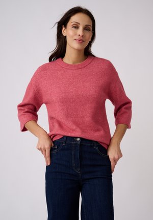 Vrouw met donker haar die een roze trui met lange mouwen en een blauwe spijkerbroek draagt, staand met handen bij de zakken tegen een effen achtergrond.