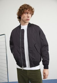 Giubbotto bomber nero con chiusura a zip, polsini e orlo a costine. Logo distintivo sul petto, indossato sopra una maglietta bianca e pantaloni in velluto a coste color oliva.