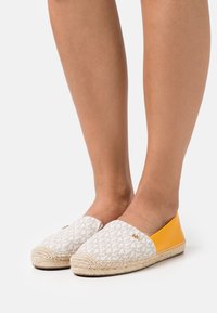 Gambe che indossano espadrillas slip-on con parte frontale in tessuto monogram beige e pannello in pelle gialla brillante sul retro, su sfondo bianco.