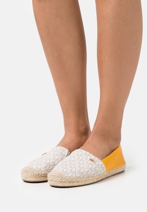 Jambes portant des espadrilles à enfiler avec un avant en tissu monogramme beige et un panneau arrière en cuir jaune vif sur fond blanc.