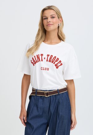Femme blonde portant un t-shirt blanc Saint-Tropez Club rentré dans un pantalon bleu taille haute avec une ceinture marron.