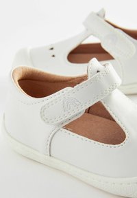 Białe skórzane buty z designem w kształcie T, miękkim, jasnobeżowym wnętrzem, okrągłymi wycięciami po bokach oraz białymi gumowymi podeszwami.