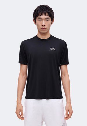 Jonge man die een zwarte EA7 Emporio Armani sport-T-shirt met korte mouwen en witte shorts draagt tegen een effen lichte achtergrond.